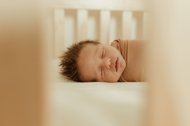 A baby sleeps in a crib, his face visible through crib slats. Denver newborn lifestyle photographer